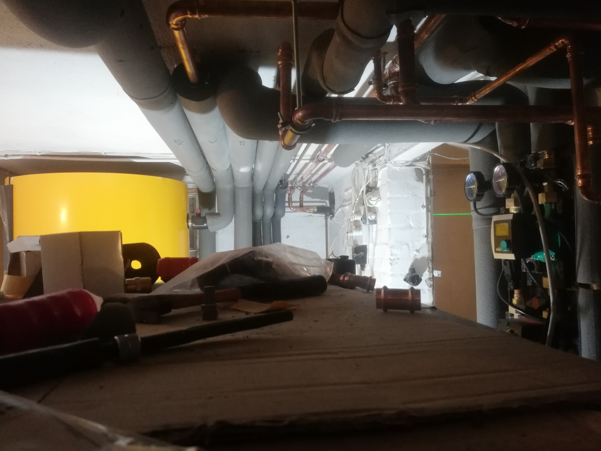 Widok z dołu na instalację centralnego ogrzewania z żółtym zbiornikiem, rurami miedzianymi i szarymi, oraz narzędziami i elementami instalacyjnymi na pierwszym planie, ukazujący etap prac...