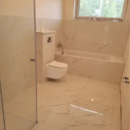 Wykończona łazienka z białymi płytkami imitującymi marmur, wisząca toaleta, zabudowana wanna, kabina prysznicowa ze szklanymi drzwiami i oknem z widokiem na zieleń.