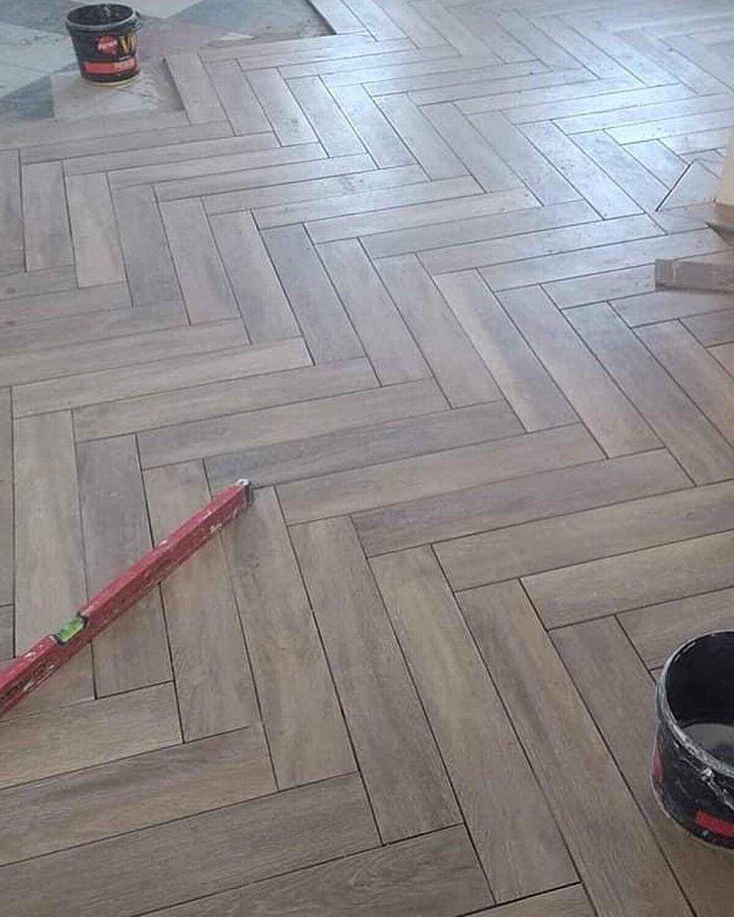 Układanie podłogi z płytek imitujących drewno w jodełkę, widoczna poziomica i wiaderko z zaprawą, etap prac wykończeniowych.