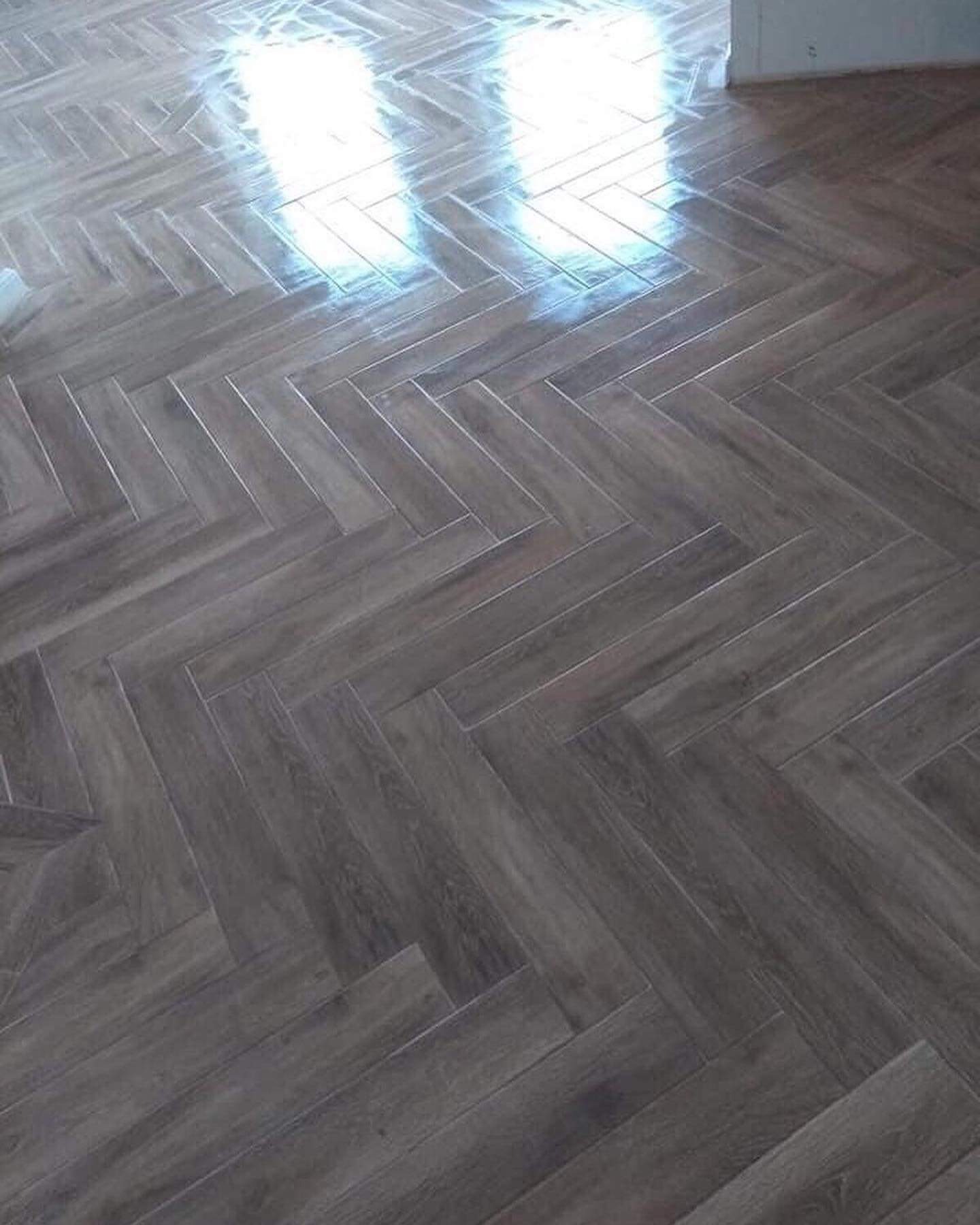 Ułożona w jodełkę, błyszcząca, szara podłoga z płytek imitujących drewno, widoczne odbicie światła.