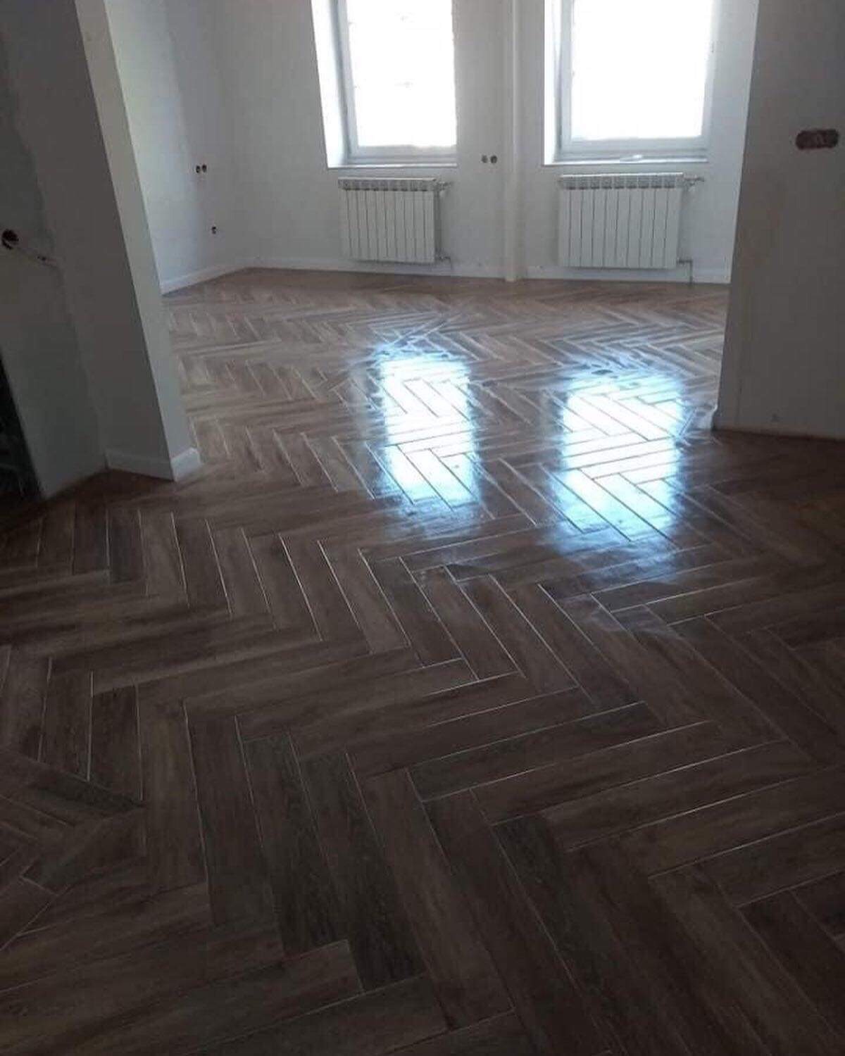 Nowo ułożona, ciemna, błyszcząca podłoga z parkietu w jodełkę w pustym, jasnym pokoju z dwoma oknami i grzejnikami.