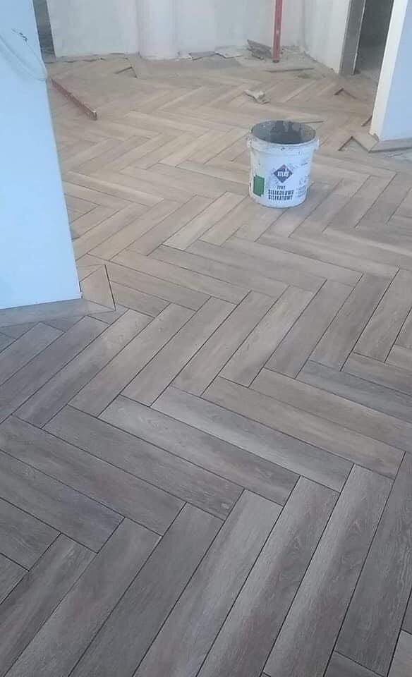 Ułożona w jodełkę podłoga z płytek imitujących drewno w odcieniach szarości, z widocznym wiaderkiem po kleju Atlas na powierzchni, w tle niedokończone ściany i podłoga.