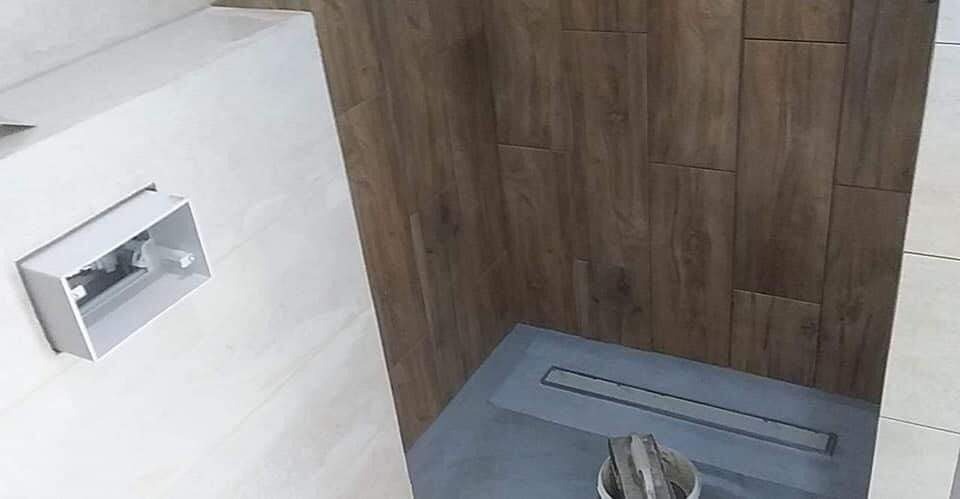 Nowoczesna łazienka w trakcie wykańczania: płytki imitujące drewno na ścianie, szary brodzik z odpływem liniowym, białe płytki i niezabudowana puszka elektryczna.