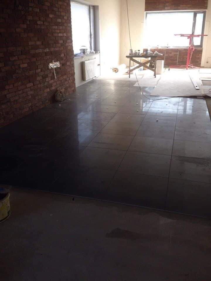 Wnętrze w trakcie remontu: ciemne płytki podłogowe ułożone na dużej powierzchni, fragment ściany z cegły, widoczne narzędzia i materiały budowlane, okna z widokiem na zewnątrz.