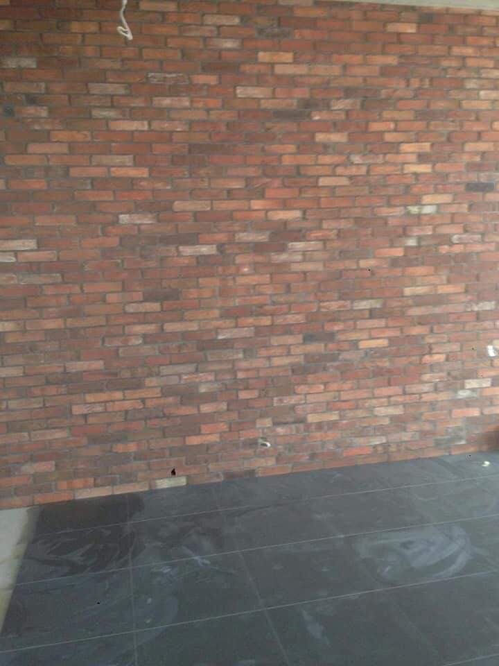 Ściana z cegły klinkierowej w odcieniach czerwieni i brązu, położona na podłodze z ciemnych, połyskliwych płytek ceramicznych w trakcie prac wykończeniowych.