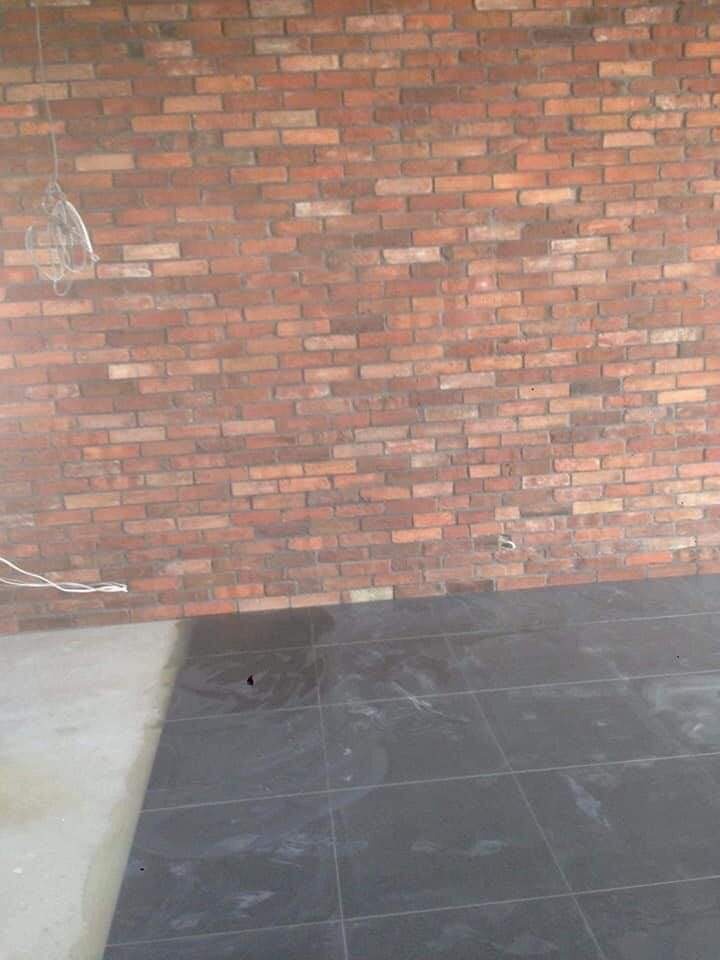 Ceglana ściana z widocznymi przewodami elektrycznymi i fragmentem ułożonej, ciemnej podłogi z płytek.