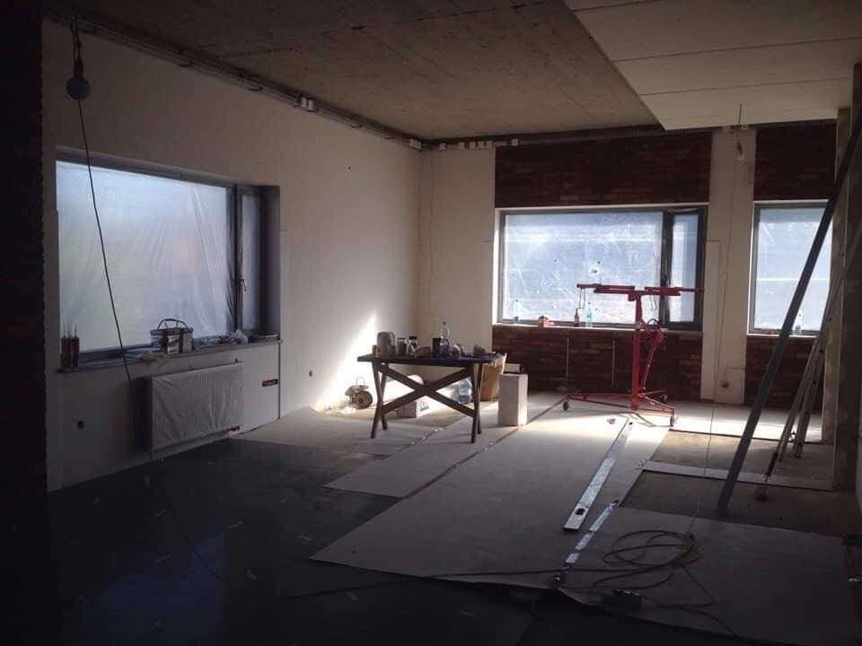 Pokój w trakcie prac wykończeniowych: zabezpieczone folią okna, ściany przygotowane pod malowanie, widoczny podnośnik do płyt gipsowo-kartonowych, narzędzia na stoliku i płyty na podłodze.