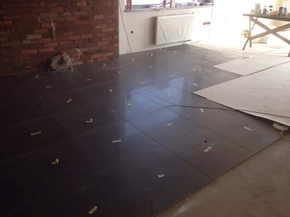 Ułożone ciemne płytki podłogowe z taśmą maskującą na łączeniach, widoczny fragment ściany z cegły i biała ściana z grzejnikiem, fragment stołu warsztatowego w tle.