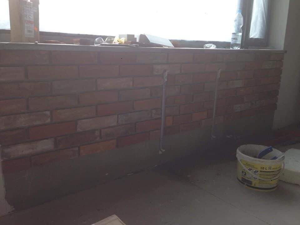 Fragment ściany z cegły klinkierowej pod oknem, widoczne rurki instalacyjne wystające ze ściany, wiadro z tworzywa sztucznego na podłodze, etap prac wykończeniowych.