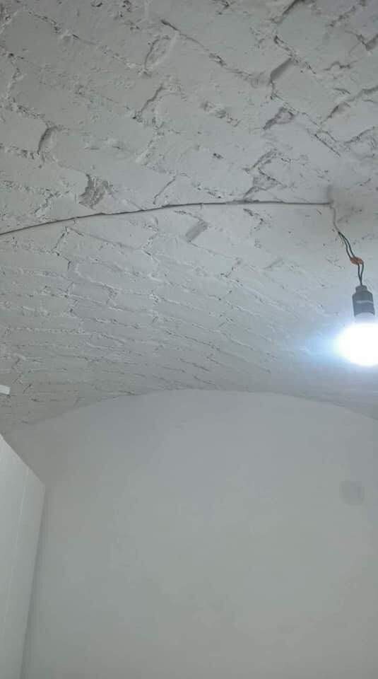 Wnętrze z łukowym sufitem z cegły pomalowanym na biało, widoczna żarówka na kablu i gładka biała ściana.