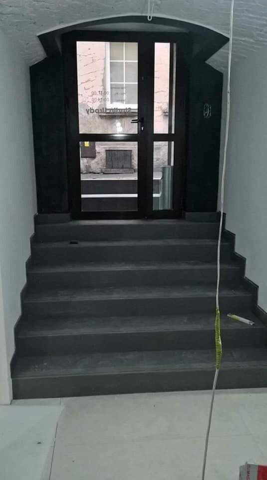 Szare schody prowadzące do ciemnych, przeszklonych drzwi wewnątrz budynku z widocznymi elementami elewacji zewnętrznej przez szybę, prawdopodobnie po remoncie, z widocznym przewodem i taśmą...