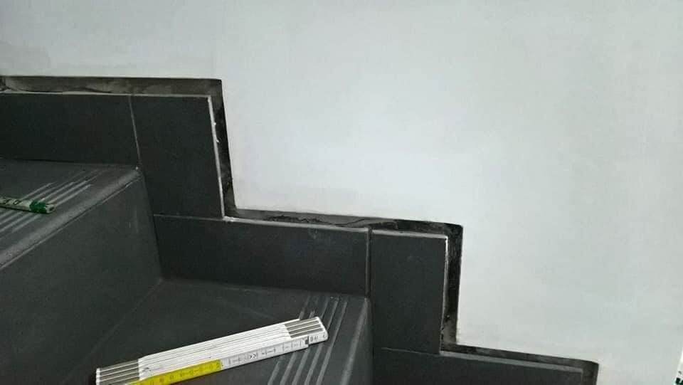 Fragment schodów z ciemnymi płytkami, widoczna nierówna fuga i biała ściana, na schodach leży rozłożona miarka stolarska.