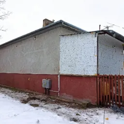 Dom w trakcie ocieplania, widoczne płyty styropianowe na jednej ze ścian, częściowo ośnieżony teren, czerwona podmurówka i szara elewacja, skrzynka elektryczna na ścianie.