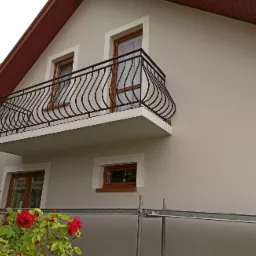 Elewacja domu po malowaniu w odcieniach beżu, z balkonem z ozdobną, czarną balustradą, okna w drewnianych ramach, widoczne rusztowanie i kwitnące czerwone róże.