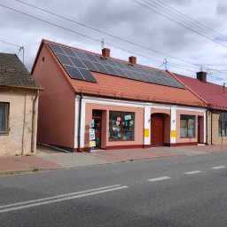 Czyste Elewacje - Odnowiony budynek z panelami słonecznymi na czerwonym dachu, widoczny z ulicy. Czerwona podmurówka i białe obramowania okien. Obok starszy budynek.