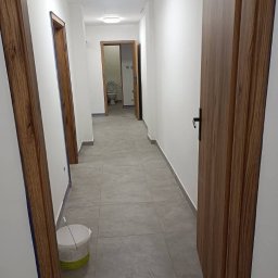 Czyste Elewacje - Wykończony korytarz z szarą podłogą i drewnianymi drzwiami. Widoczny kubeł z farbą, sugerujący świeże malowanie ścian na biało. W tle widoczna toaleta.