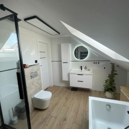 Czyste Elewacje - Nowoczesna łazienka na poddaszu z drewnopodobną podłogą, kabiną prysznicową z czarnymi ramami i okrągłym lustrem z podświetleniem LED. Uwagę zwraca skos dachu.