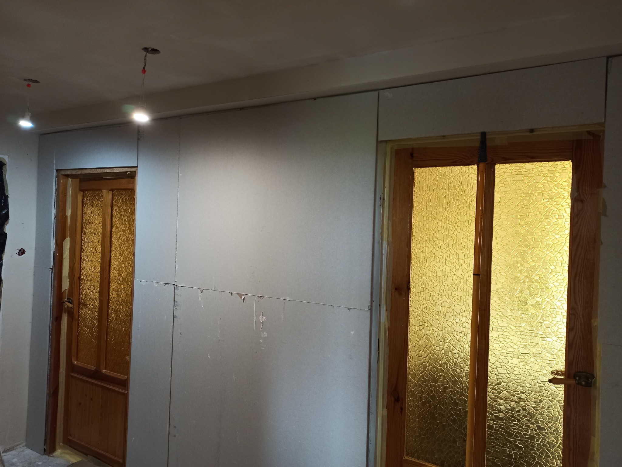 Montaż płyt kartonowo-gipsowych na ścianie w pomieszczeniu z drewnianymi drzwiami z szybami o wzorze kryształowym, widoczne niedoskonałości łączeń płyt i prowizoryczne oświetlenie.