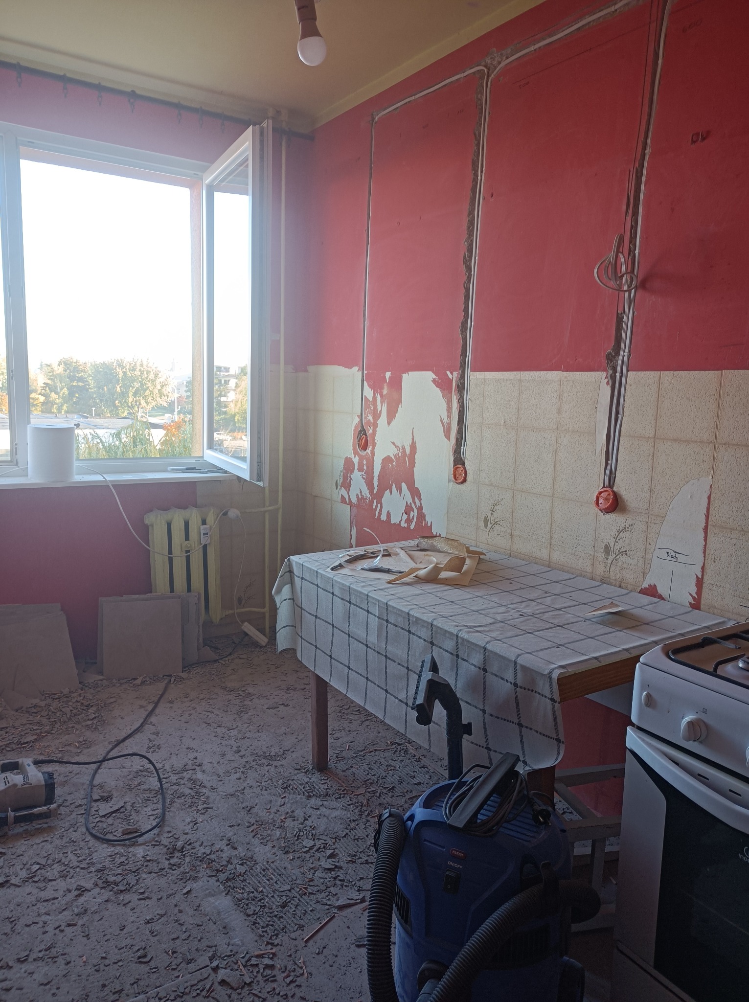 Kuchnia w trakcie demontażu starych płytek, widoczne ślady po kleju na ścianach, odkryte przewody elektryczne, narzędzia i odkurzacz przemysłowy na podłodze pokrytej gruzem.