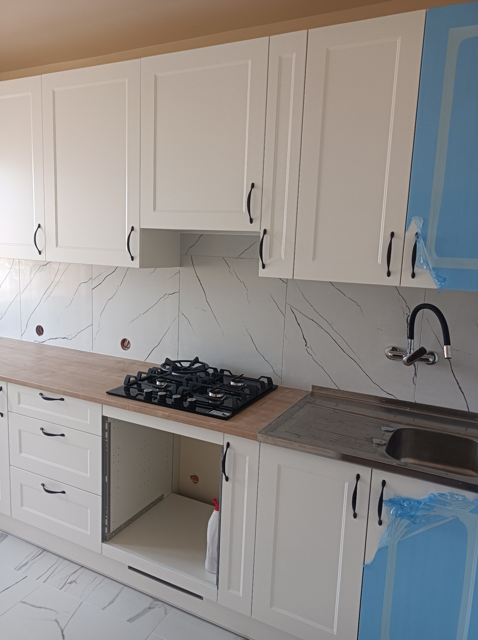 Nowoczesna biała kuchnia z marmurowymi płytkami, drewnianym blatem i czarną płytą gazową, z elementami w trakcie montażu i zabezpieczona folią ochronną.