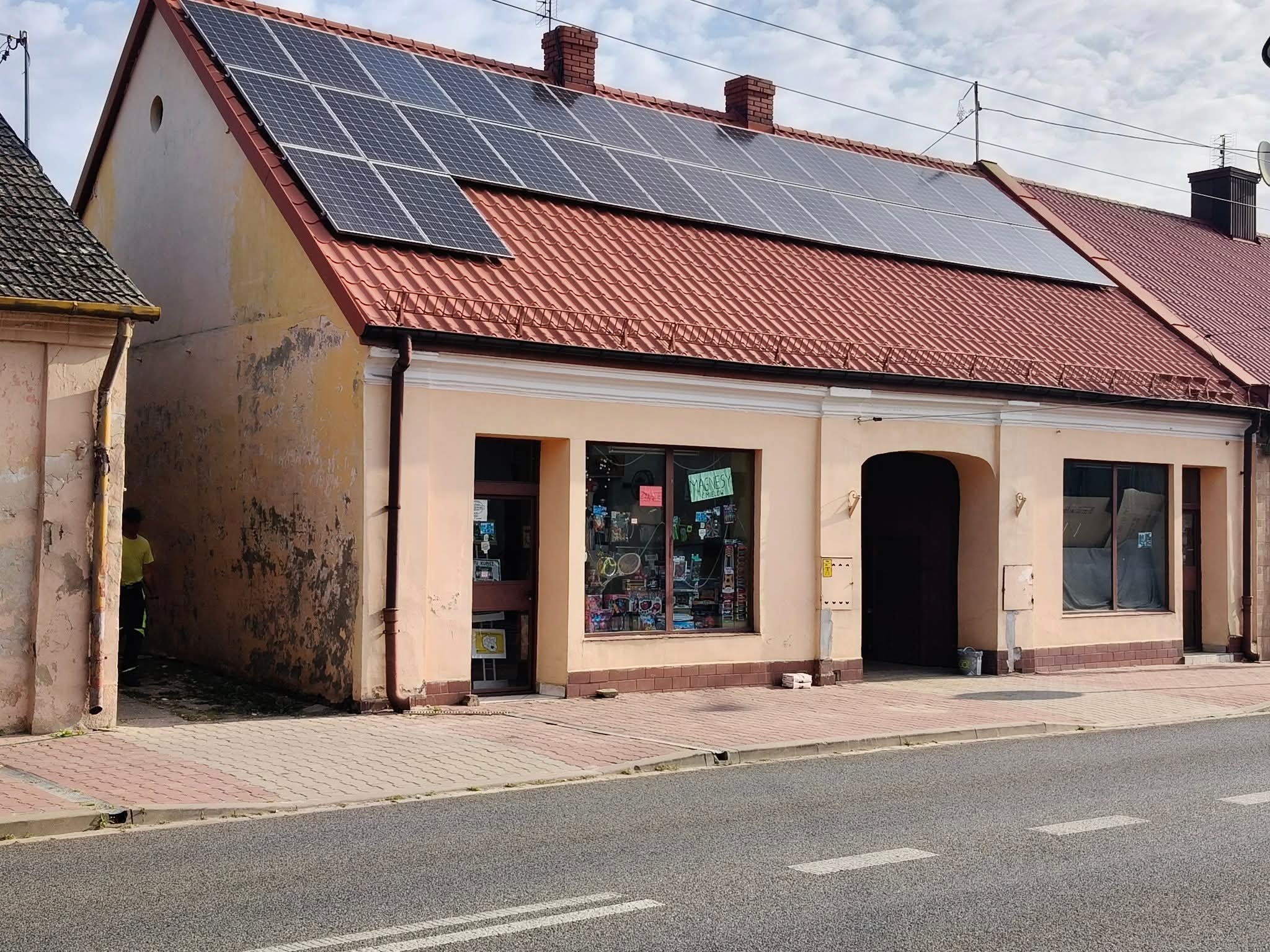 Budynek z panelami słonecznymi na czerwonym dachu, widoczne ślady zabrudzeń na elewacji. Ujęcie z poziomu ulicy, w tle błękitne niebo z chmurami.