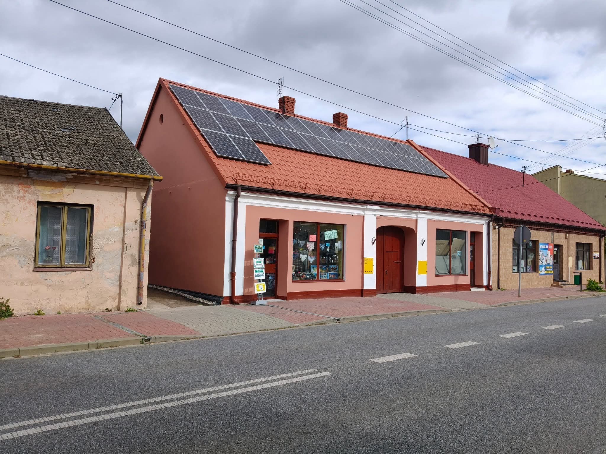 Odnowiony budynek z panelami słonecznymi na czerwonym dachu, widoczny z ulicy. Czerwona podmurówka i białe obramowania okien. Obok starszy budynek.