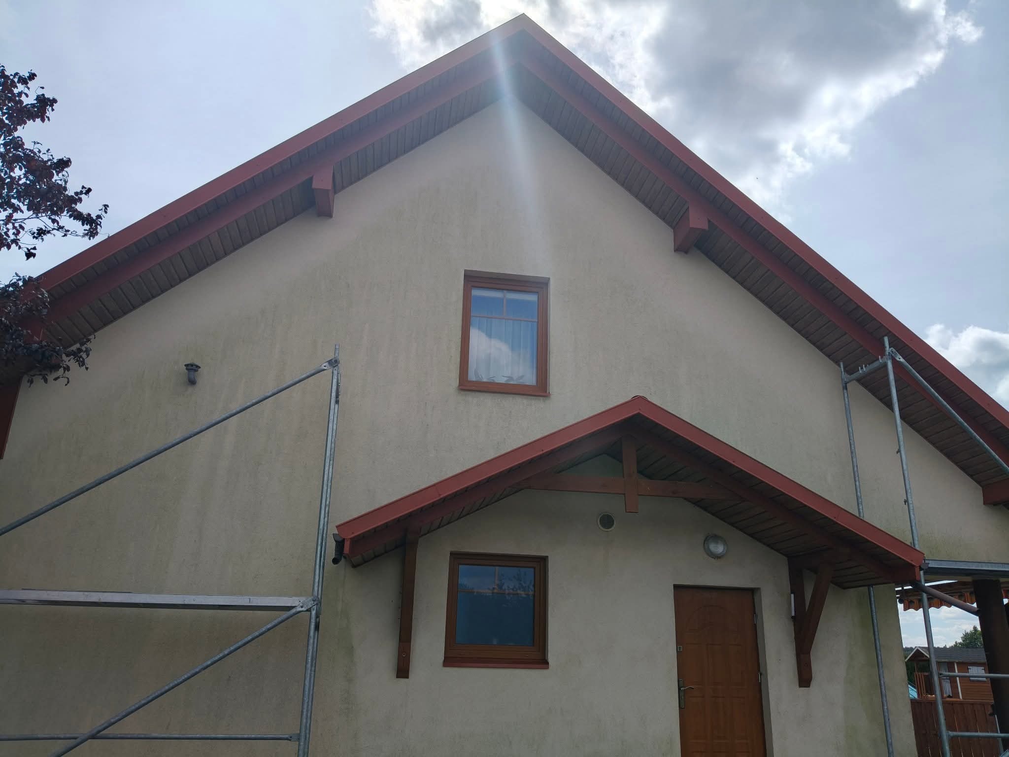 Elewacja domu jednorodzinnego w trakcie prac, widoczne rusztowanie, beżowa ściana, brązowe elementy wykończeniowe dachu i okien, pochmurne niebo.