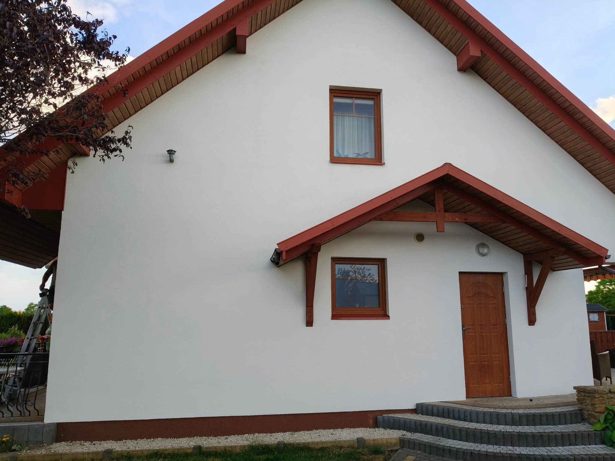 Biały dom z brązowymi elementami wykończeniowymi, widoczny z boku, z nową elewacją i schodami wejściowymi. Dom jednorodzinny po renowacji.