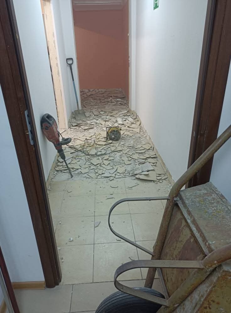 Korytarz w trakcie skuwania płytek podłogowych. Widoczny młot udarowy, łopata, gruz i taczka. Prace remontowe wewnątrz budynku.