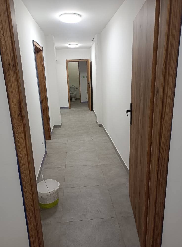 Wykończony korytarz z szarą podłogą i drewnianymi drzwiami. Widoczny kubeł z farbą, sugerujący świeże malowanie ścian na biało. W tle widoczna toaleta.