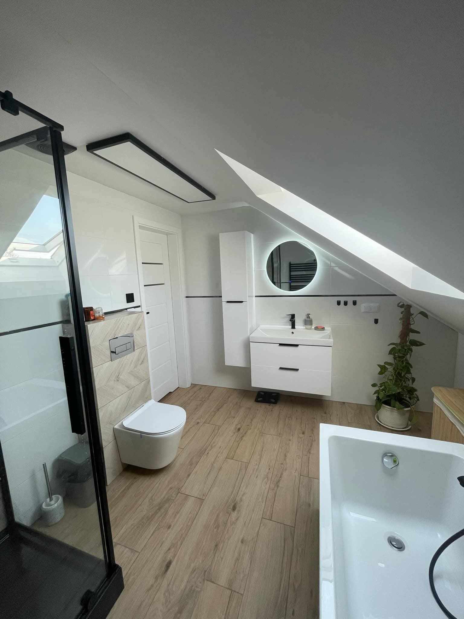 Nowoczesna łazienka na poddaszu z drewnopodobną podłogą, kabiną prysznicową z czarnymi ramami i okrągłym lustrem z podświetleniem LED. Uwagę zwraca skos dachu.
