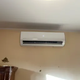 Biały klimatyzator ścienny Rotenso, z wyświetlaczem ustawionym na 30 stopni, zamontowany na beżowej ścianie nad ciemnobrązową komodą z ozdobnym, złotym elementem dekoracyjnym.