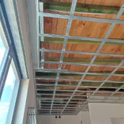 Montaż metalowej konstrukcji sufitu podwieszanego, widoczne drewniane elementy stropu i drabina Krause oparta o ścianę przy oknie.