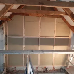 Poddasze w trakcie ocieplania, widoczne drewniane krokwie, płyty izolacyjne zamocowane na drewnianej konstrukcji, fragment drabiny i zwój kabla na podłodze, surowy stan wykończenia.