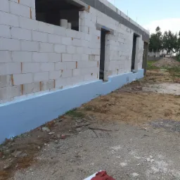 Niski mur z białych bloczków z widoczną niebieską izolacją przeciwwilgociową fundamentów, w tle niezabudowana działka i dom.