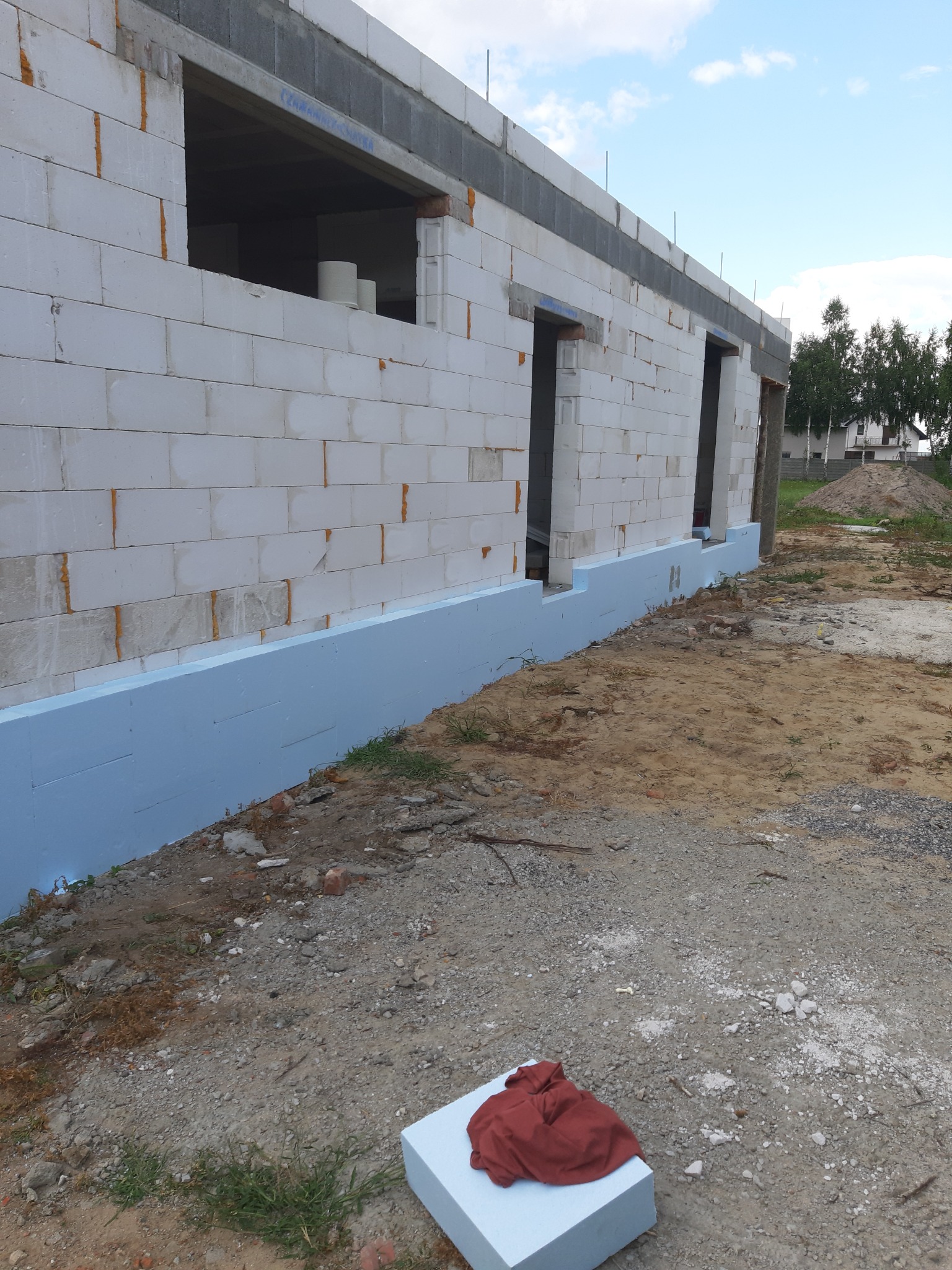Niski mur z białych bloczków z widoczną niebieską izolacją przeciwwilgociową fundamentów, w tle niezabudowana działka i dom.