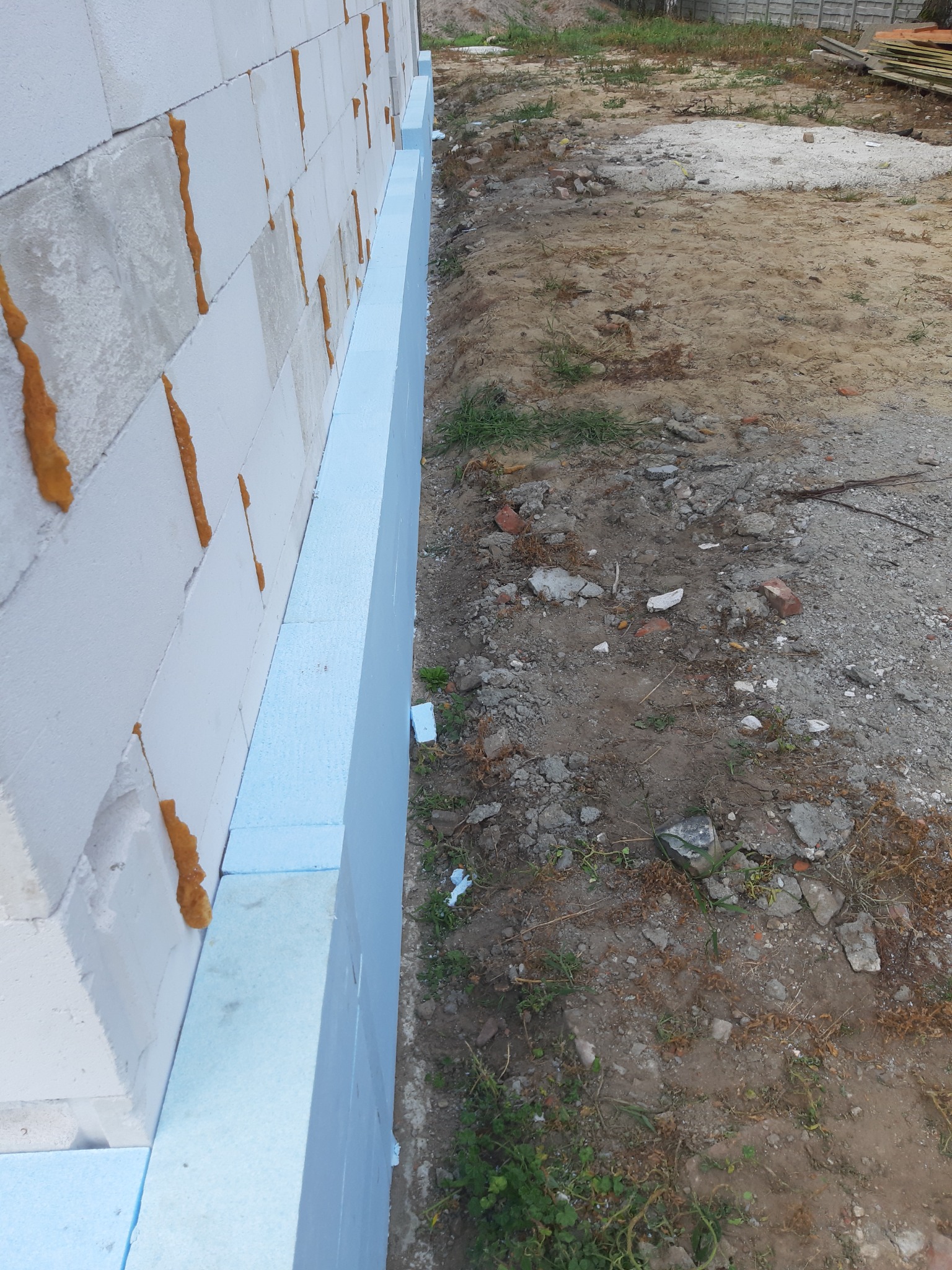 Fundament budynku z białych bloczków, częściowo ocieplony niebieskim styropianem, widoczne szczeliny wypełnione pianką montażową.