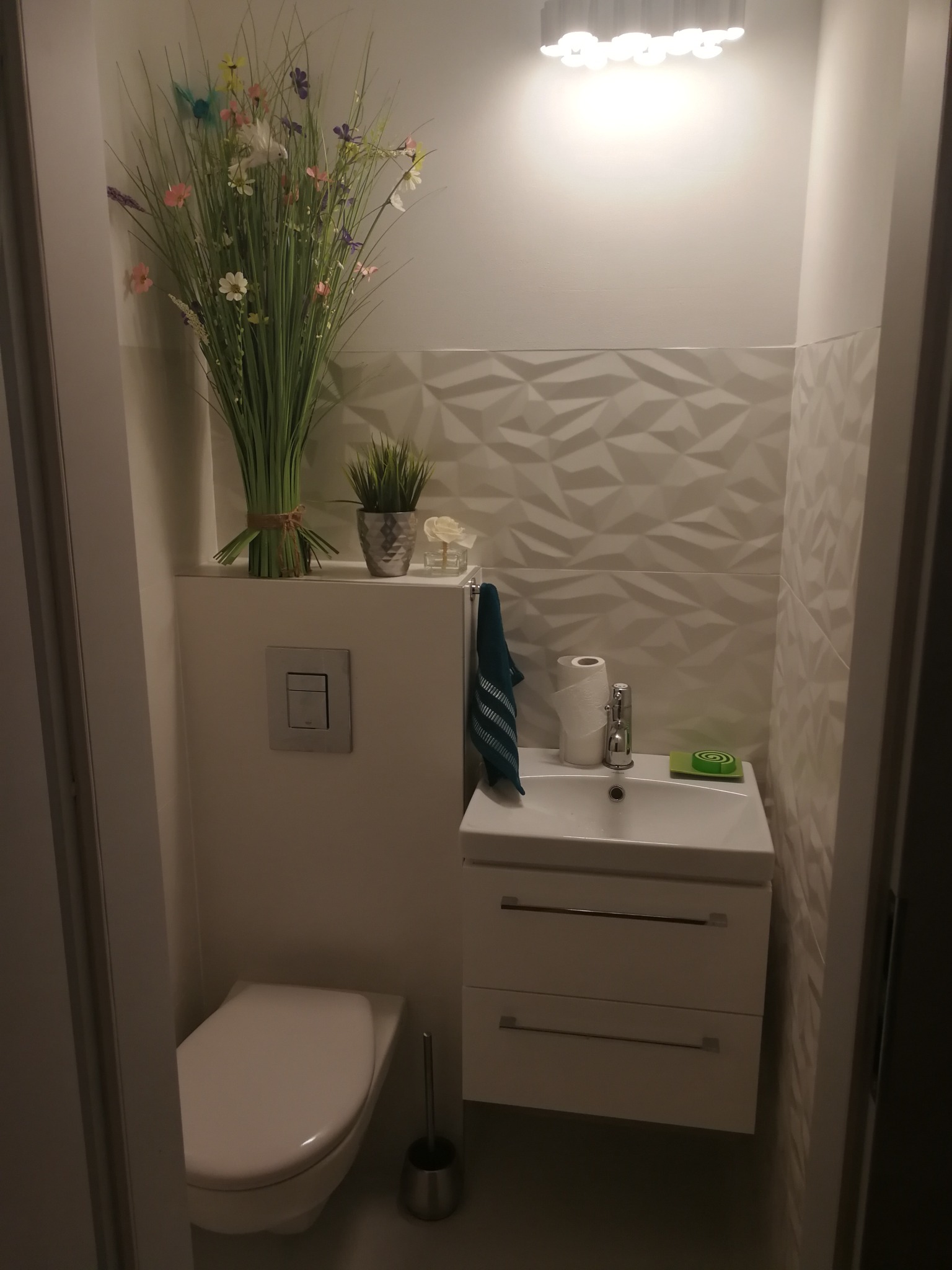 Biała, wąska łazienka z toaletą, umywalką z szafką, i dekoracyjnymi płytkami 3D na ścianie, ozdobiona bukietem sztucznych kwiatów i roślinami w doniczce.