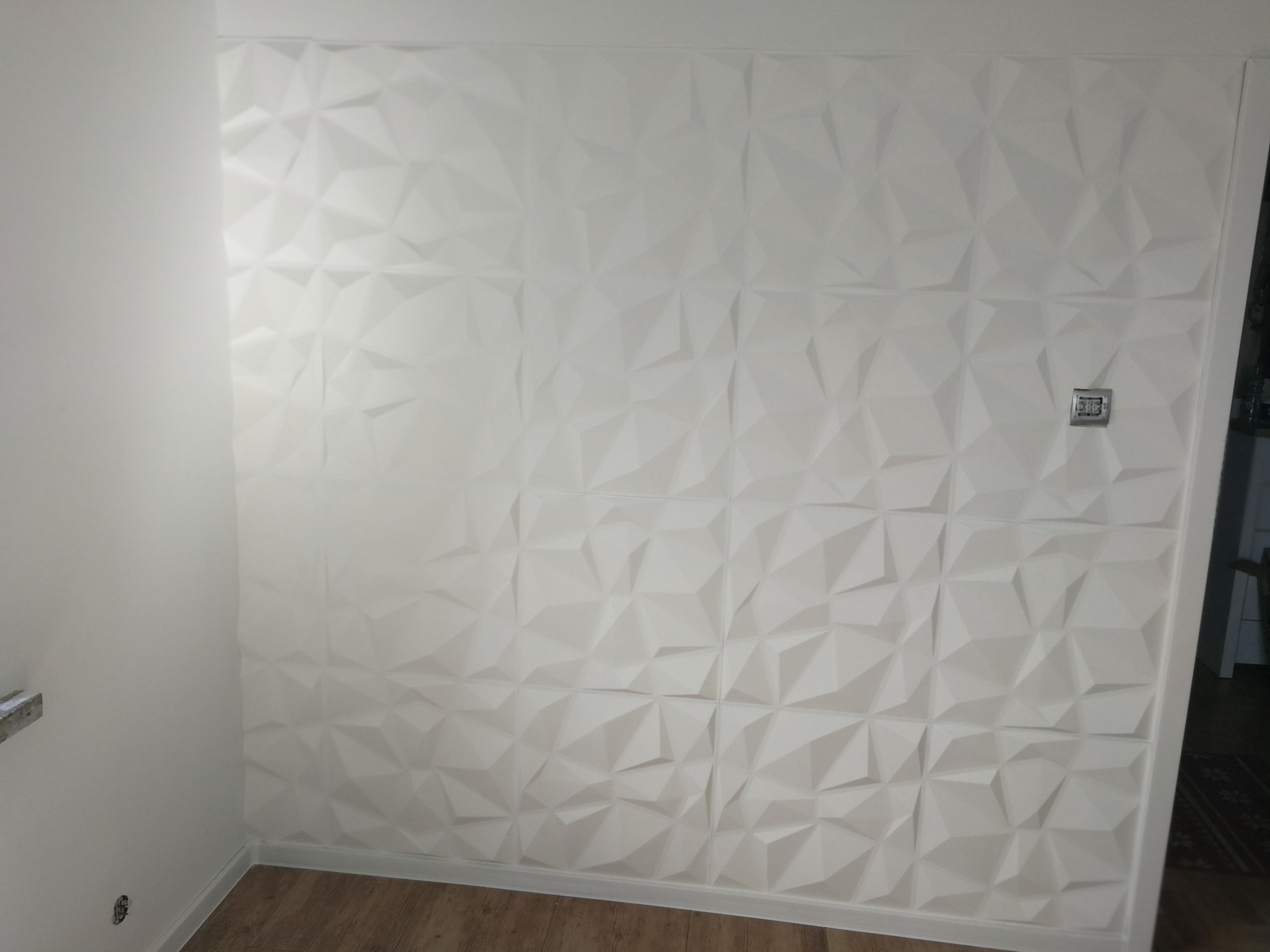 Biała ściana z trójwymiarowymi panelami dekoracyjnymi o geometrycznym wzorze, widoczny fragment drewnianej podłogi i biała listwa przypodłogowa.