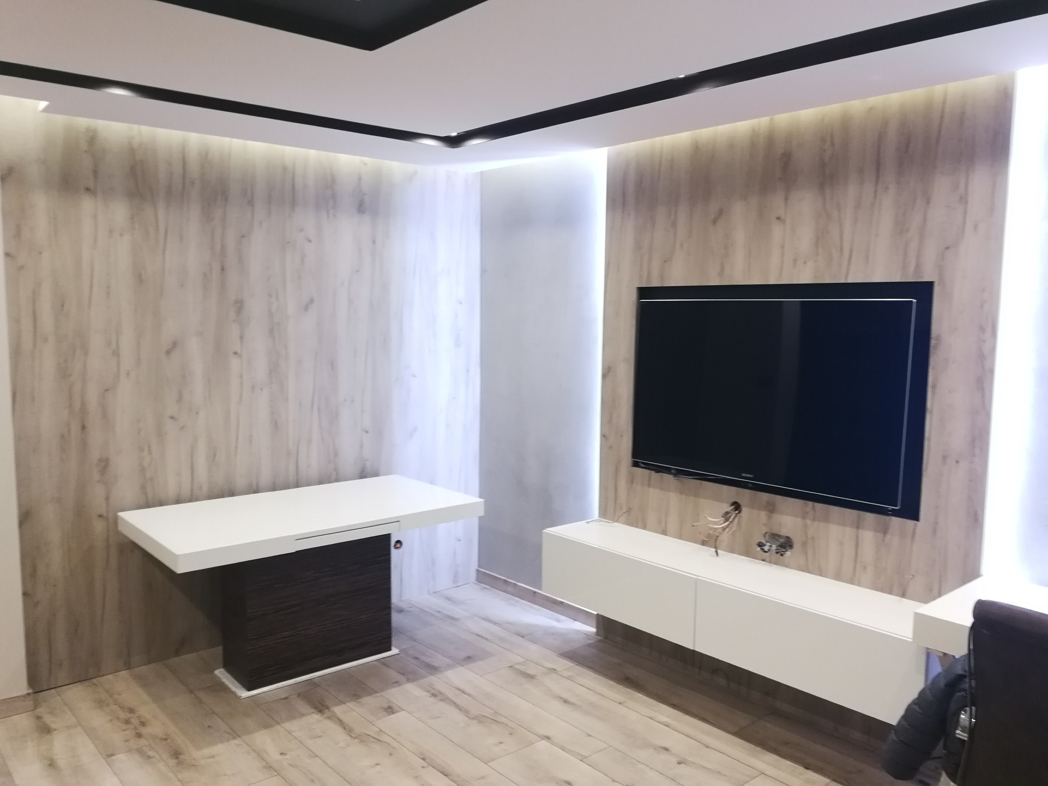 Nowoczesne wnętrze pokoju z drewnopodobnymi panelami ściennymi, białymi meblami i czarnym telewizorem na ścianie.
