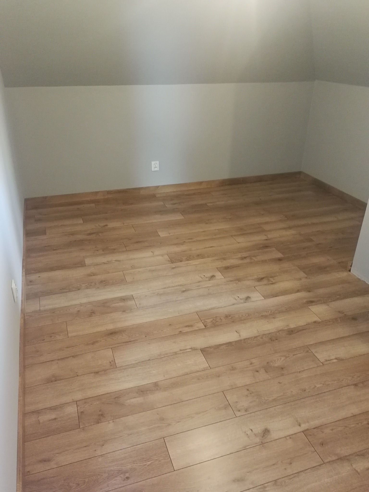 Pokój na poddaszu z jasnymi panelami podłogowymi ułożonymi wzdłuż dłuższego boku pomieszczenia, szare ściany i białe listwy przypodłogowe, widoczne gniazdko elektryczne na ścianie.