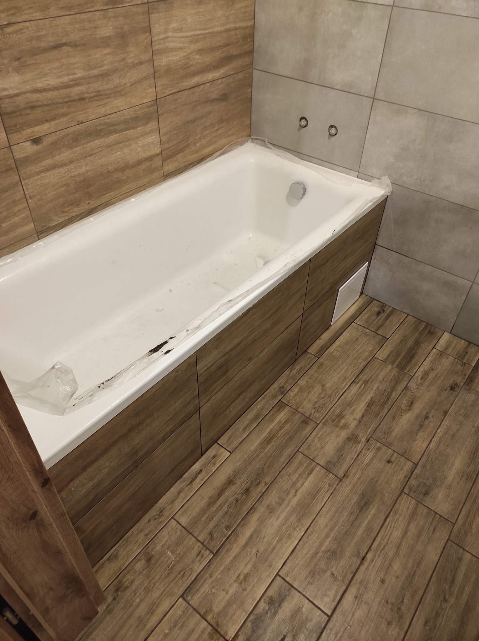 Wanna obudowana panelami imitującymi drewno, zabezpieczona folią, z widocznymi przyłączami wodnymi na ścianie wyłożonej szarymi płytkami oraz podłogą z drewnopodobnych paneli ułożonych w jodełkę.