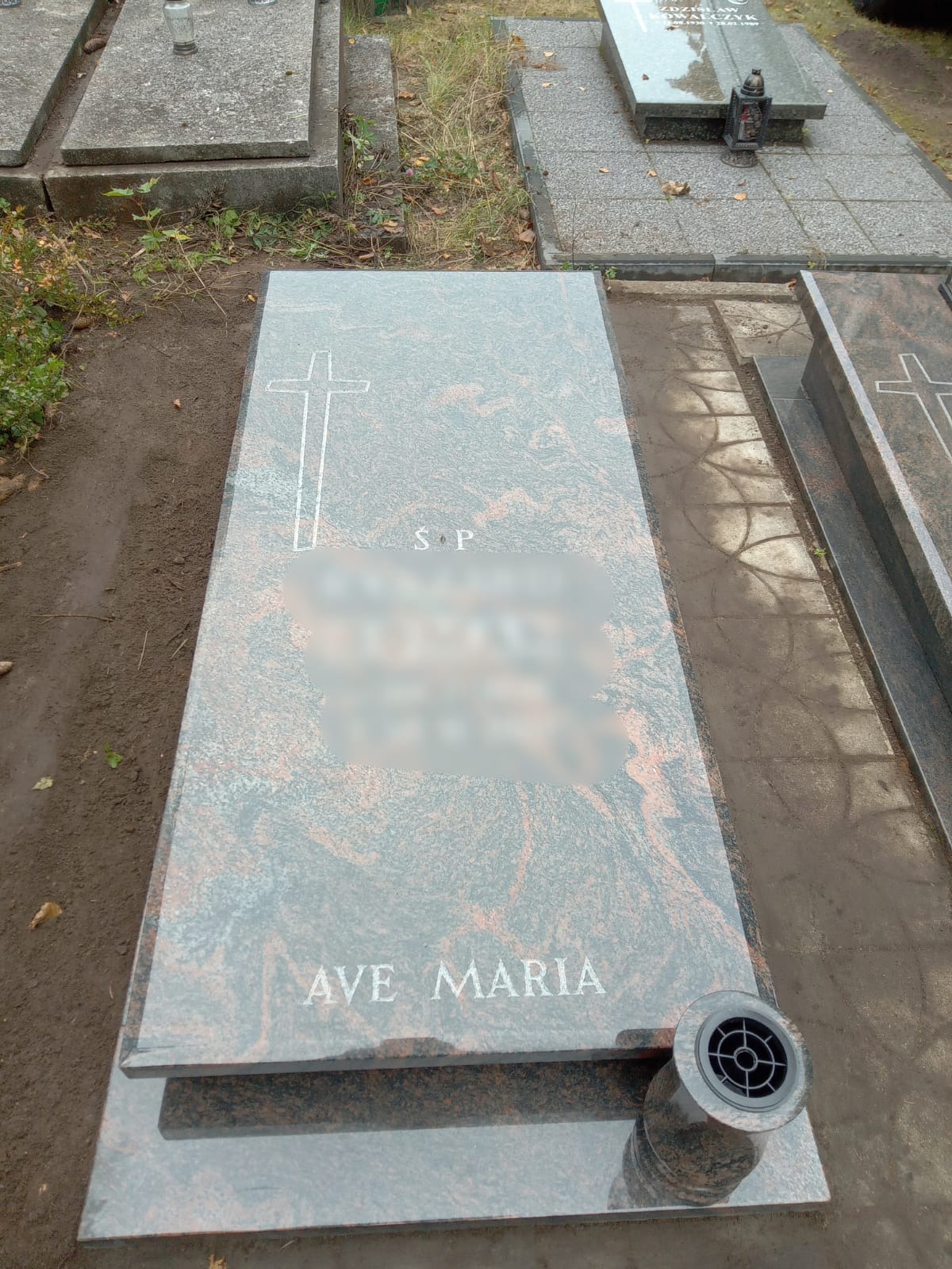 Nowy, granitowy pomnik w Kołobrzegu z wygrawerowanym krzyżem i napisem 'Ave Maria', umieszczony na cmentarzu, z widocznym wazonem na kwiaty.