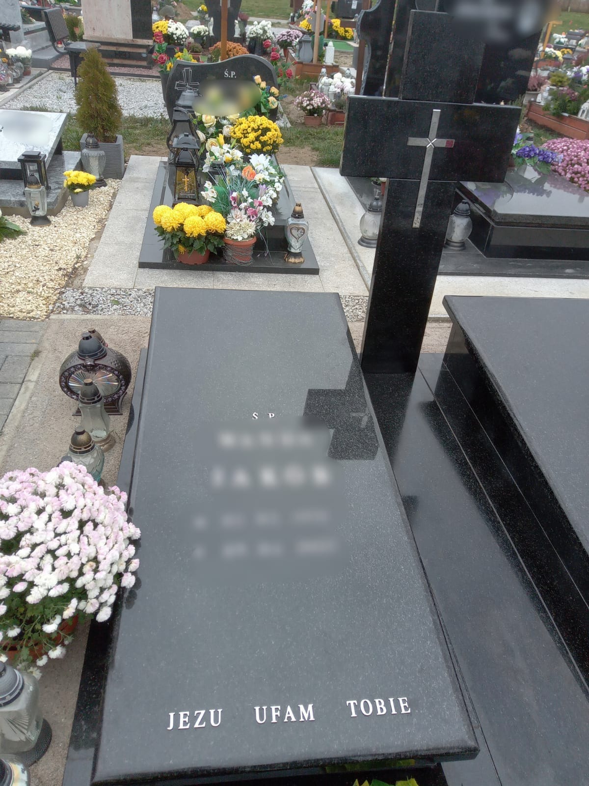 Czarny, polerowany nagrobek z napisem 'Jezu Ufam Tobie' w srebrnych literach, otoczony kwiatami i lampionami na cmentarzu.