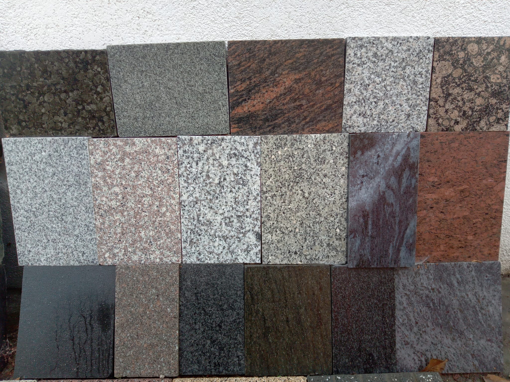 Ekspozycja próbek różnorodnych płyt kamiennych o kwadratowym kształcie i zróżnicowanej kolorystyce, od jasnych szarości po ciemne brązy i czerwienie, prezentująca tekstury i wzory kamienia.