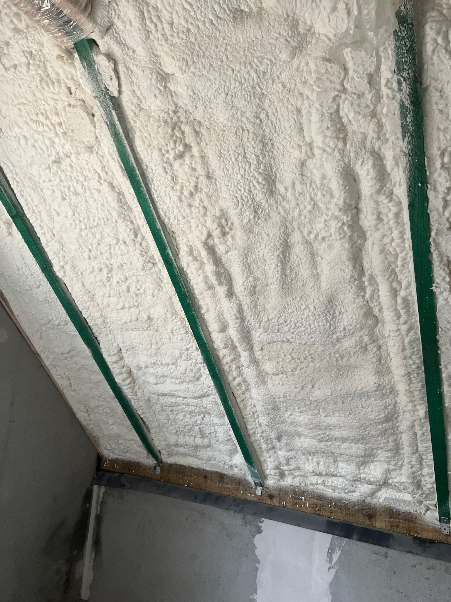 Poddasze ocieplone białą pianą natryskową, widoczne zielone elementy konstrukcyjne i fragment ściany z surowego betonu.