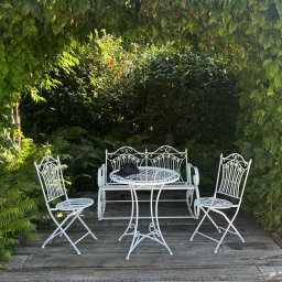 STARK ZAKŁAD USŁUGOWO-HANDLOWY EWA WRÓBEL - Biały zestaw mebli ogrodowych z metalu, składający się z ławki, stołu i dwóch krzeseł, umieszczony na drewnianym podeście, otoczony zielenią pnączy i paproci, z czarną czapką leżącą na stole.