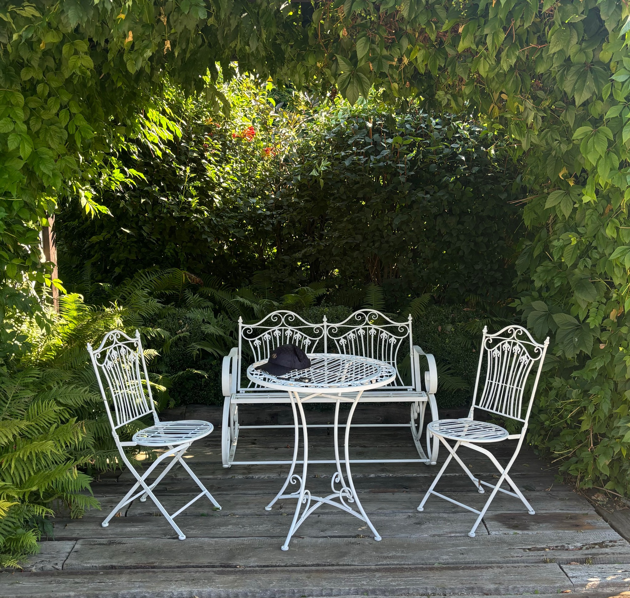 Biały zestaw mebli ogrodowych z metalu, składający się z ławki, stołu i dwóch krzeseł, umieszczony na drewnianym podeście, otoczony zielenią pnączy i paproci, z czarną czapką leżącą na stole.