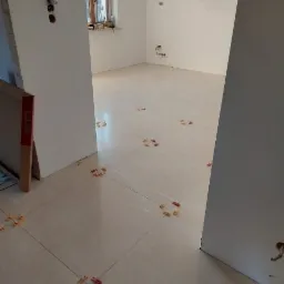 Układanie jasnych płytek podłogowych w pomieszczeniu z użyciem krzyżyków dystansowych, widoczne fragmenty okna i instalacji elektrycznej.