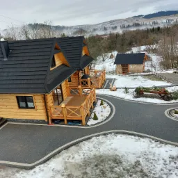 Widok z góry na posesję z drewnianymi domkami w górskim stylu, z czarną kostką brukową tworzącą ścieżki i podjazdy, otoczoną śnieżnym krajobrazem i zalesionymi wzgórzami w tle.